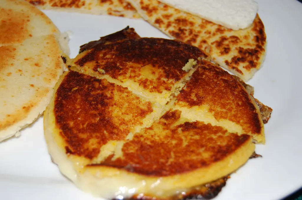 Golden seared corn arepas on a white plate, one cut into wedges with melted cheese