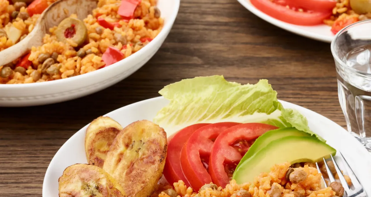 Plate of orange rice with pigeon peas and olives, with fried plantains, avocado, and tomato