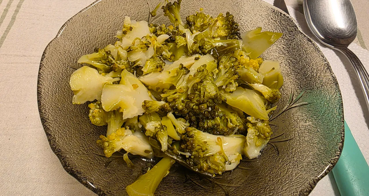 Cooked broccoli florets and stems in a textured glass bowl, next to a teal-handled spoon