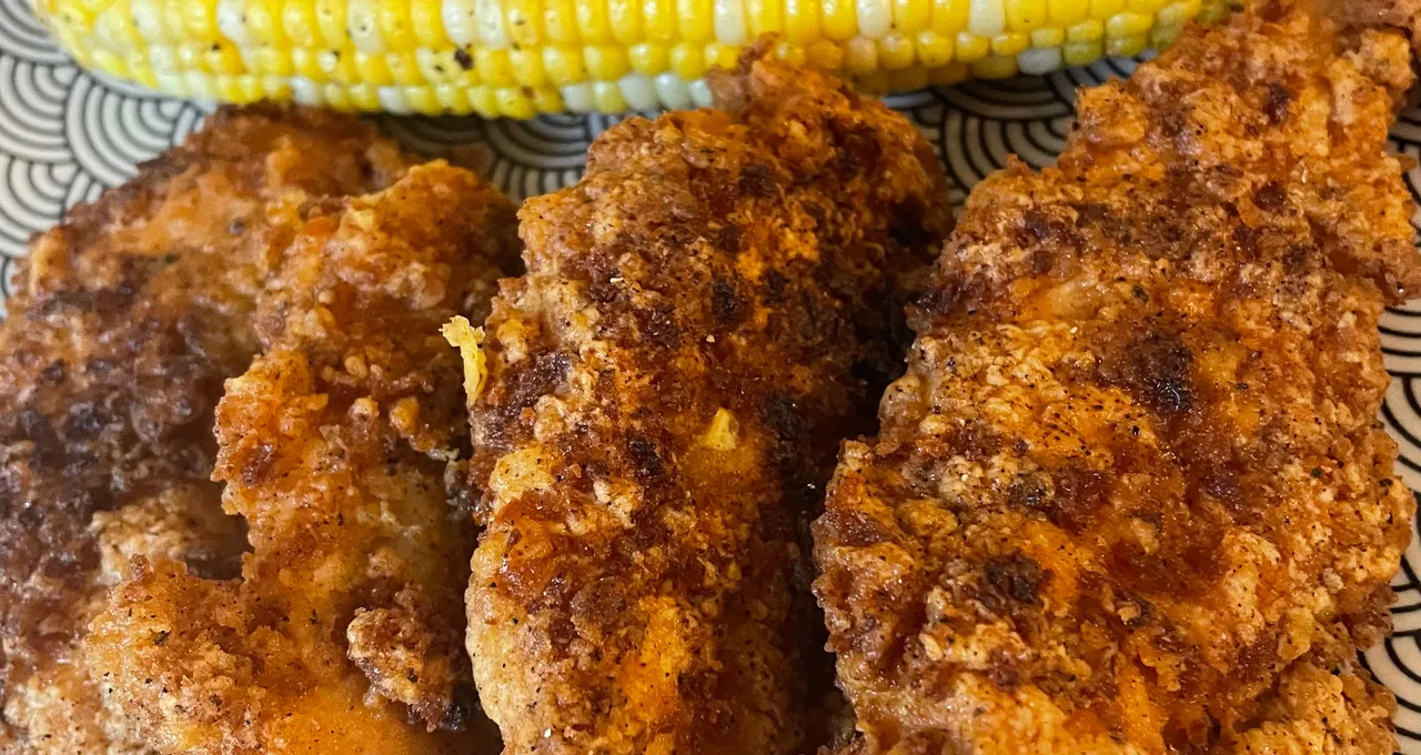 Three fried chicken tenders and a cob of corn on a black-and-white patterned plate