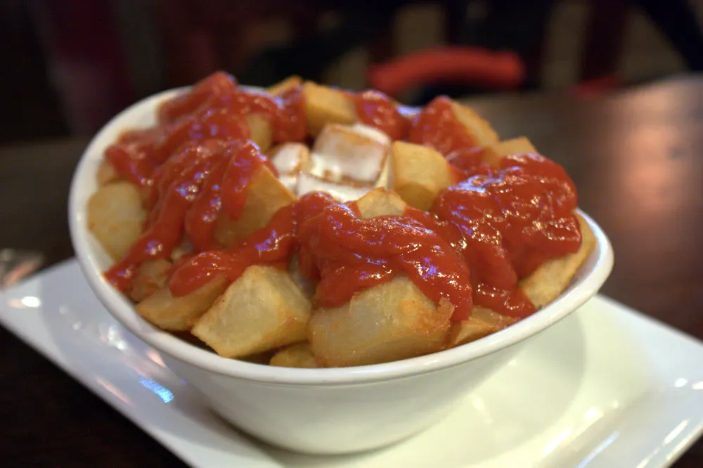 Cubed fried potatoes in a white bowl, red brava sauce and aioli on top